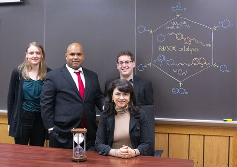 Group picture of the researchers with Christina White seated at table and others standing in front of chalk board with the reaction drawn on the board.