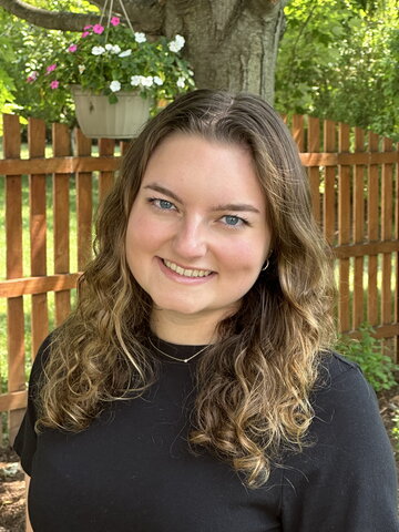 Outdoor portrait of Amalia standing in a grassy area with a light brown picket fence in background 