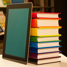 Photo of a tablet leaning against a stack of books, that are "pride" colors, on a table