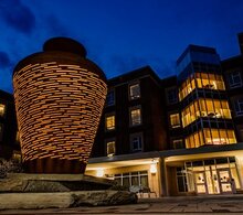 An art installation featuring a metal basket with light from within shining through the basket cracks sits in front of Wassaja Hall at night. 