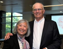 Susan Morisato and Tom Remec standing side by side in room with a window and video screen in the background.