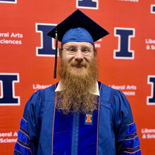 Zane Thornburg stands in graduation regalia in front of orange backdrop