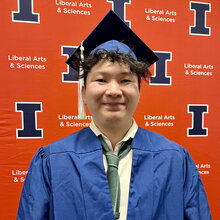 Zipeng Shen stands in graduation regalia in front of orange backdrop