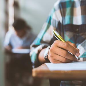 Photo partially showing a student at a desk writing with a pen