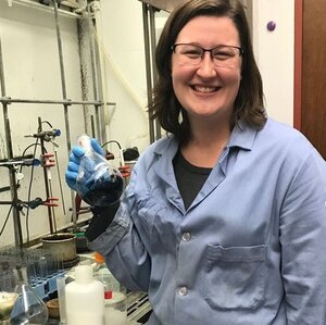 Photo of Amanda East in a blue lab coat in the lab holding a beaker with liquid 