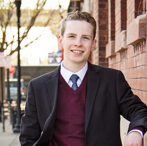 Portrait of Evan Dray standing on a sidewalk next to a brick building in a suit jacket, sweater, collared shirt and tie. 