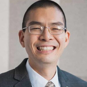 Head shot of Grant Hisao in a dark suit coat, shirt and tie on a gray background