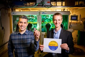 Beniamin Zahiri holds a small battery between two fingers standing in a lab next to Paul Braun who is holding a diagram on a white sheet of paper that illustrates their design.