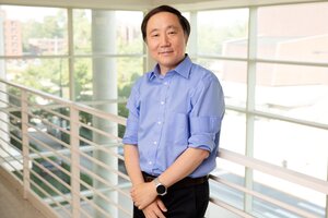 Portrait of Yi Lu in a building on campus, standing on a balcony that overlooks a wall of large windows with other campus buildings in the background.