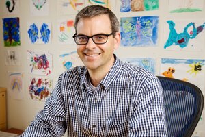 Portrait of Martin Burke sitting in a chair in his office with pictures of drawings on the wall behind him.