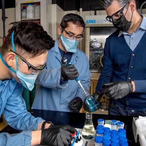 Three researchers wearing masks and rubber gloves stand around a table as one researcher fills a vial.