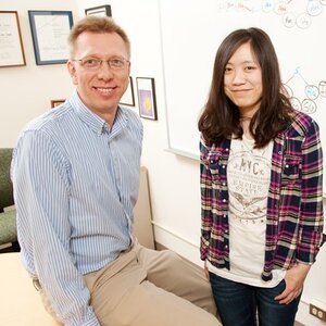 Portrait of Prof. Wilfred van der Donk, on left, sitting on a desk with graduate student Weixin Tang standing to the right. Framed documents and a wipe off board are on walls behind them.