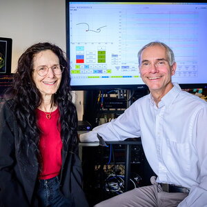 Carla Scaletti and Martin Gruebele sit in front of two large computer screens that show data sonification data and charts