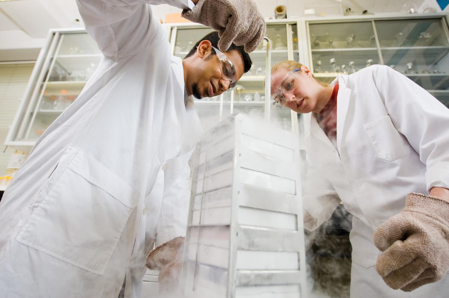 Two chemistry students in white lab coats work in a lab looking over stacked trays.
