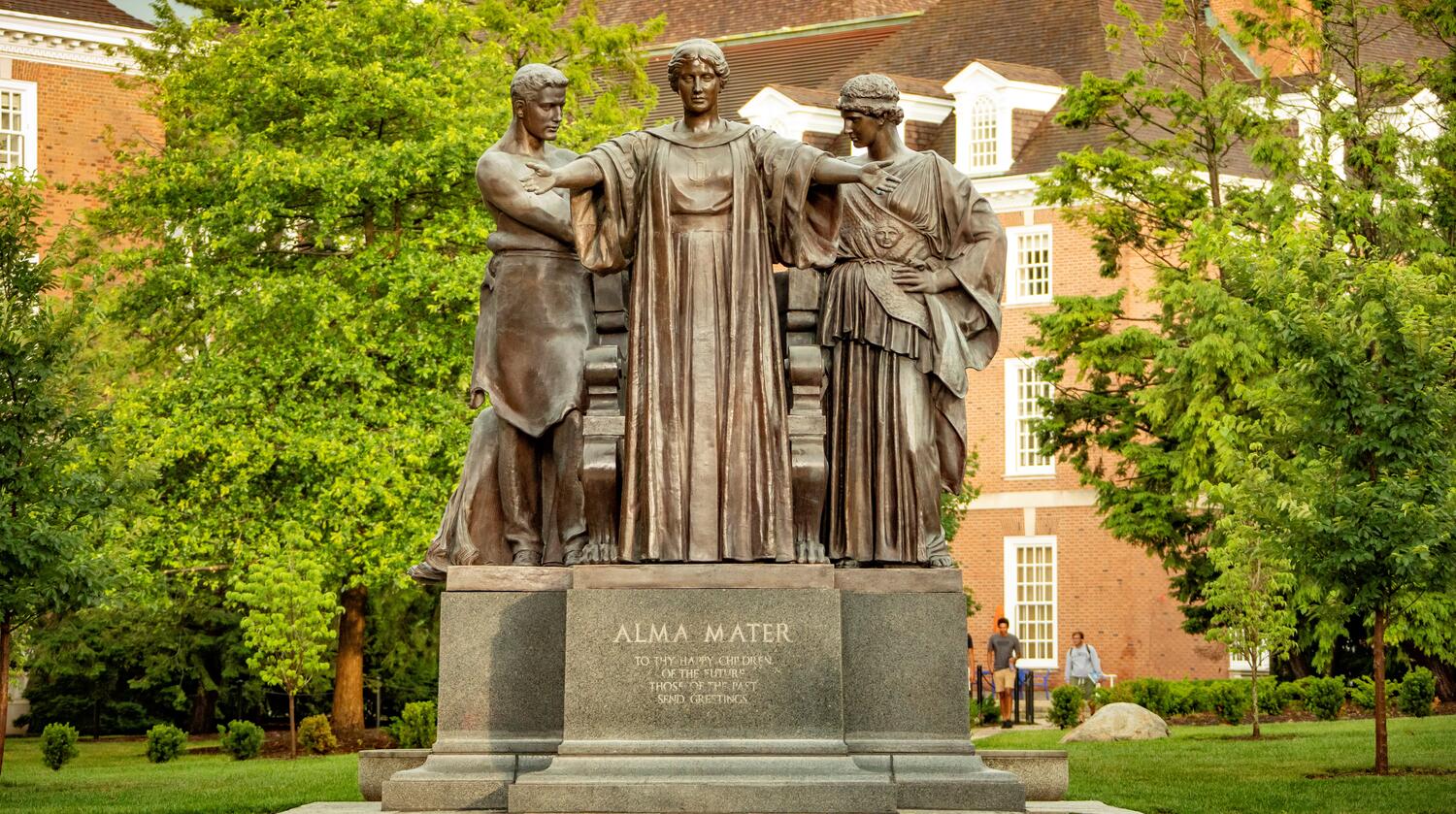 Photo of the Alma Mater Statue on campus during the summer with green trees and nearby brick buildings in the background
