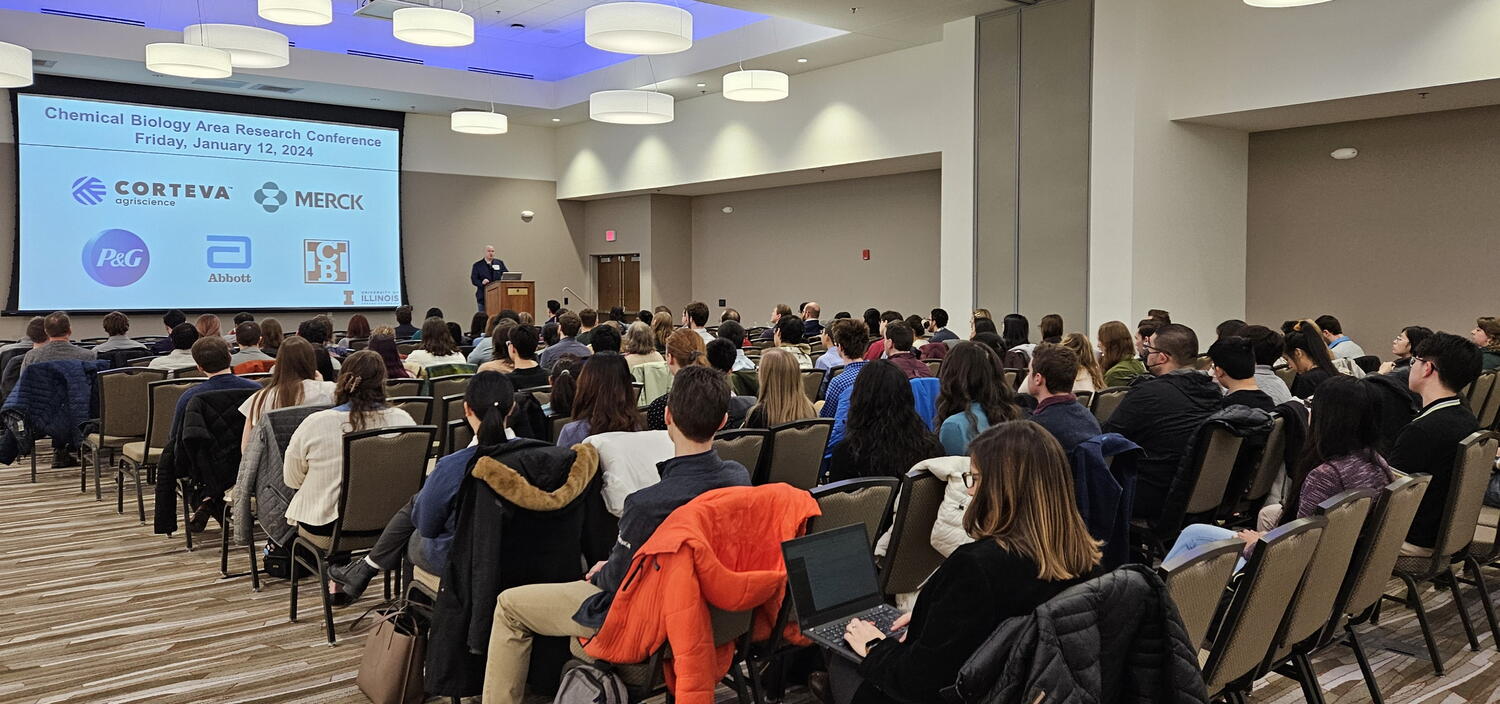 Audience sits in chairs listening to speaker at the front of a room at podium with a large presentation screen.