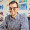 Portrait of Martin Burke sitting in a chair in his office with pictures of drawings on the wall behind him.