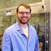 Andrew Zahrt stands in a blue lab coat in a chemistry lab with equipment in the background.