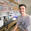 Graduate student Jason Cournoyer sits on a chair in a lab next to a bench and shelves filled with chemistry supplies/equipment.