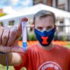 A student standing outdoors on the UIUC campus with a COVID testing tent site in the background holds a tube with blue liquid at the bottom, like the tubes used for collecting COVID saliva samples.