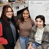 Student researchers stand and sit side by side in a lab with a wipe off board behind them with chemical equations from their research.