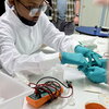 A middle school student wearing a white lab coat and turquoise lab gloves sits at a table working on building a solar cell.