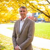 Portrait of Paul Hergenrother standing outdoors with yellow fall colored trees and a pathyway in the background.