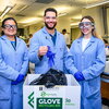 Three lab instructors wearing blue coats and PPE stand side by side in a teaching lab in Chem Annex and the center instructor is dropping a blue glove in a glove recycling box.