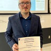 Francis Alcorn stands holding the Klemperer Award certificate in a classroom with a large overhead screen in the background